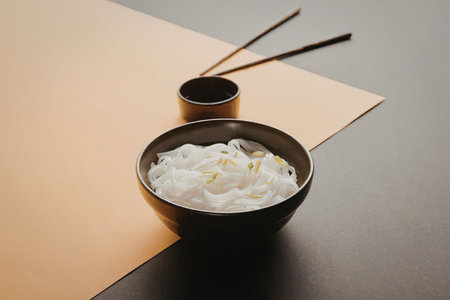 A black bowl filled with white noodles and green herbs on a yellow and gray surface with chopsticks...