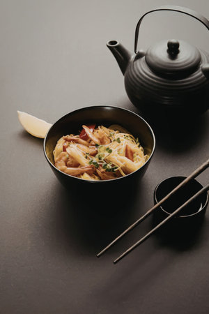A bowl of noodles with vegetables and herbs, accompanied by chopsticks, a small bowl, and a teapot...