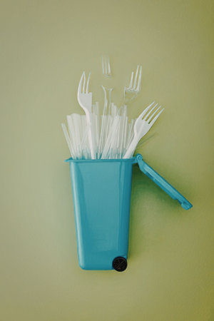 A small blue trash can with a black wheel and handle overflowing with white plastic forks and other...