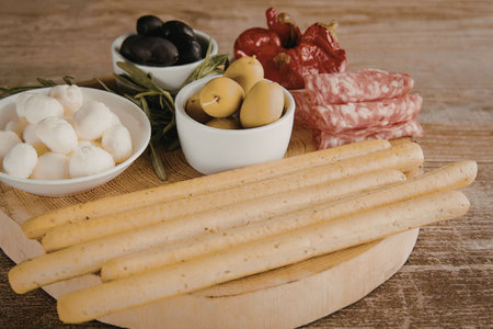 A wooden board with breadsticks, mozzarella balls, olives, and cured meatsの写真素材