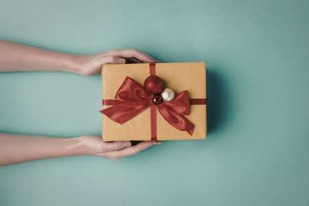 A person holding a gift box wrapped in yellow paper with a red ribbon and bow, and decorated with...