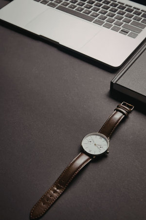 A brown leather watch with a white face is placed on a dark surface.