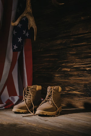 A pair of brown work boots with yellow soles and laces, placed on a wooden floor in front of a wooden wall and an American flag.の写真素材