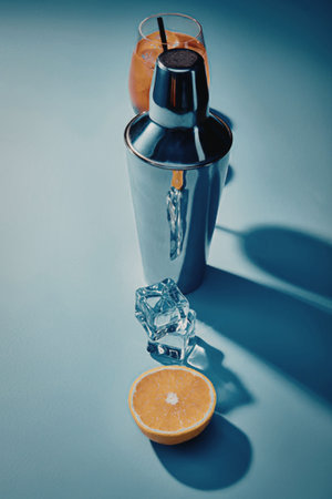 A cocktail shaker with a glass of orange juice on top, an ice cube falling into it, and a half...