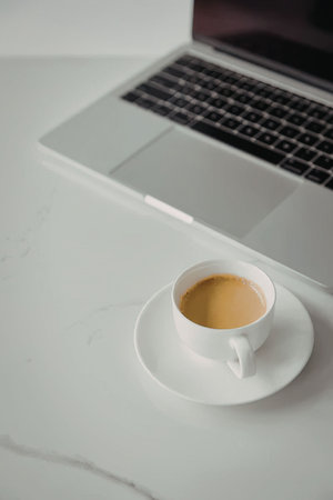 A white coffee cup on a saucer sits on a white marble desk next to a silver laptop.