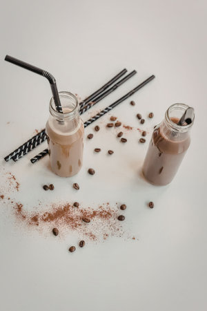 Two glass bottles of iced coffee drinks with straws, coffee beans, and powder on a white surfaceの写真素材