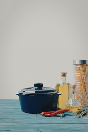 A blue pot with a lid, two bottles of oil, red chili peppers, and a jar on a blue wooden table.の写真素材