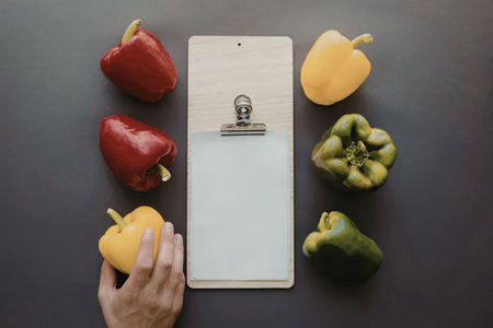 A hand holding a yellow bell pepper next to a clipboard surrounded by red, green, and yellow bell...