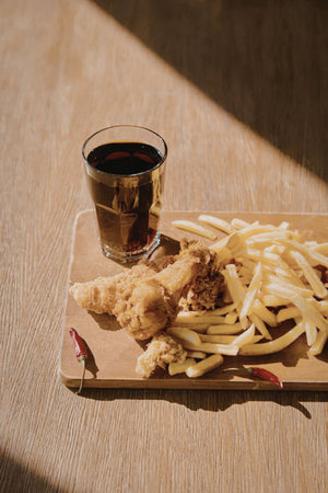 A wooden plate with fried chicken, french fries, and a glass of soda on a wooden table.