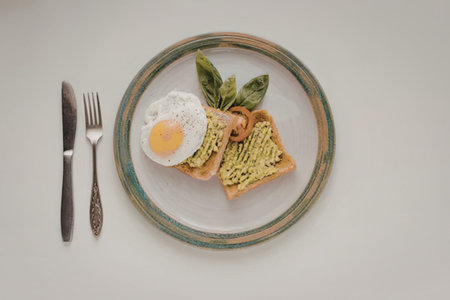 A plate with avocado toast, a fried egg, and fresh herbs, accompanied by a knife and fork.