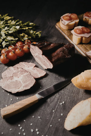An arrangement of sliced meats, bread, tomatoes, and herbs on a dark table.の写真素材