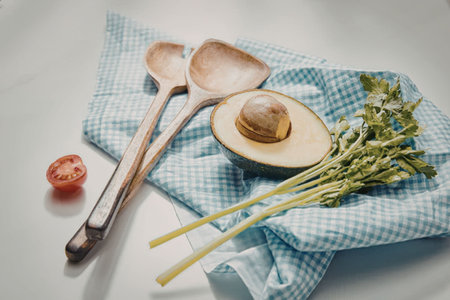 An avocado half with pit, two wooden spoons, asparagus, and a cherry tomato on a blue and white...