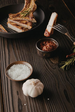 A plate of grilled meat with a fork holding a piece of bread, accompanied by a bowl of salt, a bulb of garlic, a bowl of red sauce, and some herbs on a dark wooden table.の写真素材