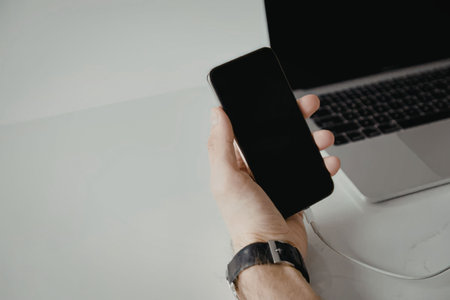 A hand holding a smartphone with a blank screen next to a laptop on a white surface.
