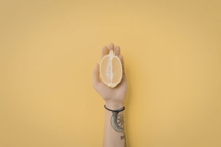 A hand with a tattoo on the forearm holding a half lemon against a bright yellow background.