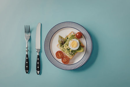 A plate of food with egg, tomatoes, and vegetables, accompanied by a fork and knife on a light blue...