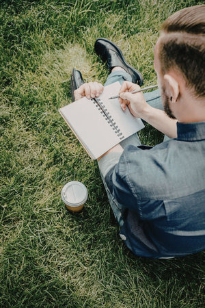 A man sits on the grass, writing in a notebook with a pencil. A coffee cup is next to him.の写真素材