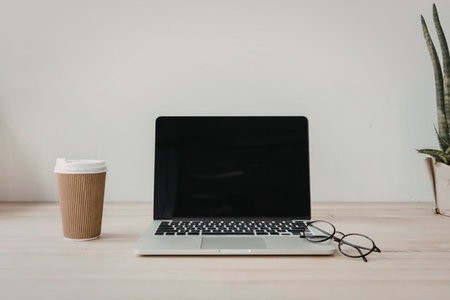 A laptop computer sits on a light-colored wooden desk next to a disposable coffee cup and a pair of...