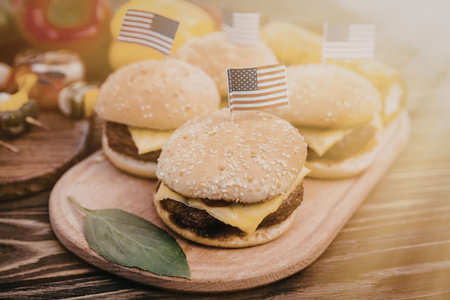 A plate of mini burgers with american flags on top, served on a wooden plate with a green leaf and other food items in the background.の写真素材