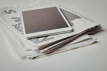 A stack of newspapers with a white tablet on top, The tablet has a blank screen and is placed on a...