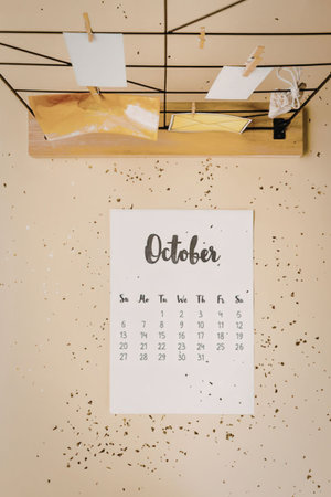 A calendar page for October hangs on a yellow wall with a wooden shelf above it, holding clothespins with paper and a small clock.の写真素材