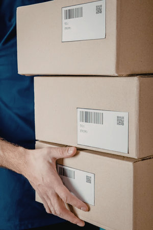 A person in a blue shirt is holding a stack of three brown cardboard boxes with barcode labels.
