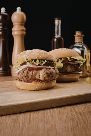 Two burgers on a wooden cutting board with condiments in the background.の写真素材
