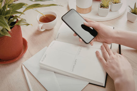 A person holding a phone and a notebook on a desk with a plant, cup of tea, and other items
