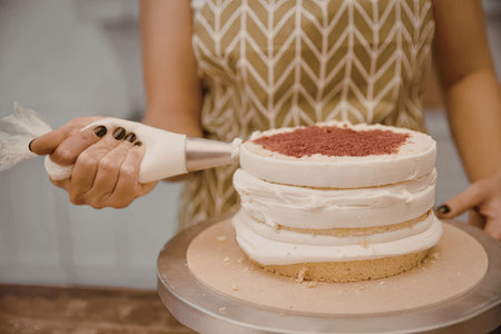 A woman in a chevron-patterned dress is shown decorating a layered cake with white frosting and red...