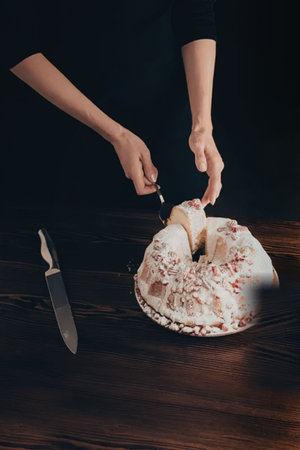 A person cutting a slice of cake with white frosting and red sprinkles on a dark wooden table.