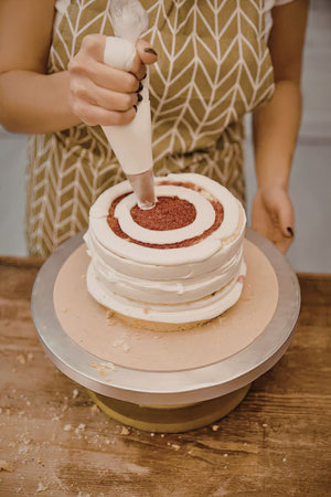 A person in a green and white apron is decorating a multi-layered cake with white frosting and red...