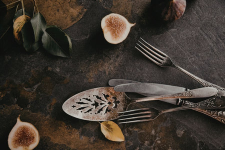 A dark and moody still life featuring sliced figs, a spoon, fork, and knife on a rustic surface.