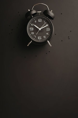 Black alarm clock with white numerals and hands on a dark background with scattered dots and ample...