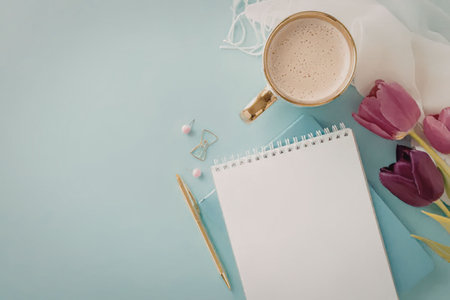 Blank notebook lies open on a blue surface accompanied by a gold pen and cup of coffee surrounded by...