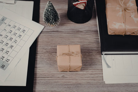 Christmas gifts and notepad on a wooden desk with a calendar and small christmas tree nearby,...