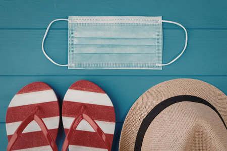 Striped flip flops and straw hat are placed beside a face mask on a blue wooden surface, symbolizing...