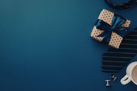 Gift wrapped in brown paper with blue ribbon placed on blue background with tie, cufflinks, and...