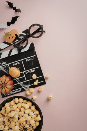 Bowl of popcorn and film clapboard decorated with pumpkins and bats on a pink background symbolizing a Halloween movie night celebration.