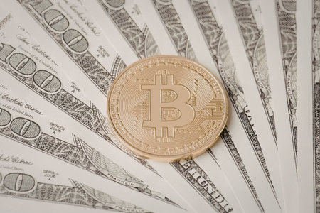 Golden bitcoin coin resting on top of a fan of usd dollar banknotes illustrating the intersection of...