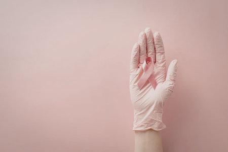 Hand wearing pink latex gloves holding a pink breast cancer awareness ribbon against a soft pink background.の写真素材