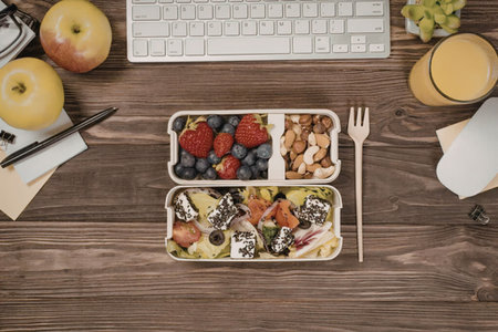 Healthy meal in lunch box with mixed greens fruits and nuts placed beside a laptop keyboard on a wooden office desk with a refreshing glass of orange juice.の写真素材