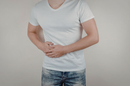 Man wearing a white t-shirt and jeans holding his abdomen in pain and experiencing discomfort with...