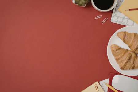 Croissants on a white plate accompanied by coffee and digital devices on a red workspace with...
