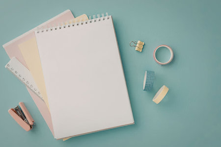Stationery supplies featuring a blank spiral notebook, colored papers, a ruler, stapler, binder clip, and decorative tapes are neatly organized on a bright blue surface.の写真素材