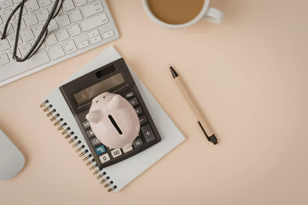 Calculator with pink piggy bank on top of spiral notebook accompanied by pen and cup of coffee on...