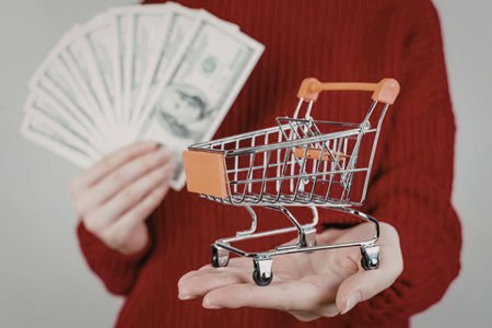 Person wearing red sweater holds miniature shopping cart and fan of usd currency notes in their...