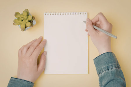 Person's hands holding a blank notepad and pen on a yellow background with a small green plant nearby, symbolizing creativity and inspiration in a simple workspace setting.の写真素材