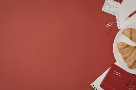 Croissants on a white plate placed on a red and white desk with notebooks and red stationery supplies creating a cozy and inviting workspace for a productive morning.の写真素材