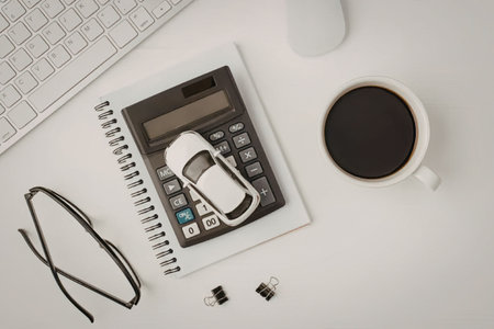 Calculator and car key on a notebook, glasses, and a coffee cup on a clean white desk, symbolizing a...