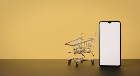 Smartphone with a blank white screen stands beside a small metal shopping cart on a dark surface against a bright yellow background, symbolizing online shopping and ecommerce.の写真素材